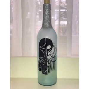Puede incluir: Una botella de vidrio esmerilado con una tapa de purpurina plateada. La botella tiene una silueta en blanco y negro de Jack Skellington y Sally de Pesadilla antes de Navidad.