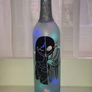May include: A frosted glass bottle with a silver glitter top and a black silhouette of Jack Skellington and Sally from the Nightmare Before Christmas. The bottle is lit with multi-colored fairy lights.