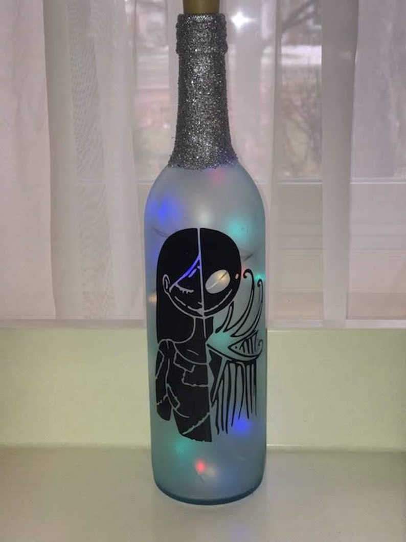 May include: A frosted glass bottle with a silver glitter top and a black silhouette of Jack Skellington and Sally from the Nightmare Before Christmas. The bottle is lit with multi-colored fairy lights.