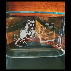 Könnte beinhalten: Eine transparente Glasbackform mit hellblauem Rand und Deckel. Die Form zeigt ein geätztes Design einer Vintage-Frau, die ein Schild mit der Aufschrift "Bitch, I AM the secret ingredient!" hält.