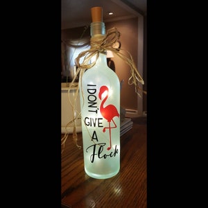 Könnte beinhalten: Eine dekorative Glasflasche mit Korken, verziert mit einer roten Flamingo-Grafik und dem schwarzen Schriftzug "I DON'T GIVE A Flock". Die Flasche ist mit kleinen Lichtern beleuchtet und mit Bast gebunden.