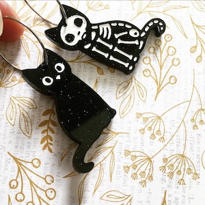 以下が含まれることがあります： キラキラ光る仕上げの黒い猫のイヤリングが2つ。左側の猫は尻尾を脚に巻き付けて座っています。右側の猫は尻尾を上げて歩いています。