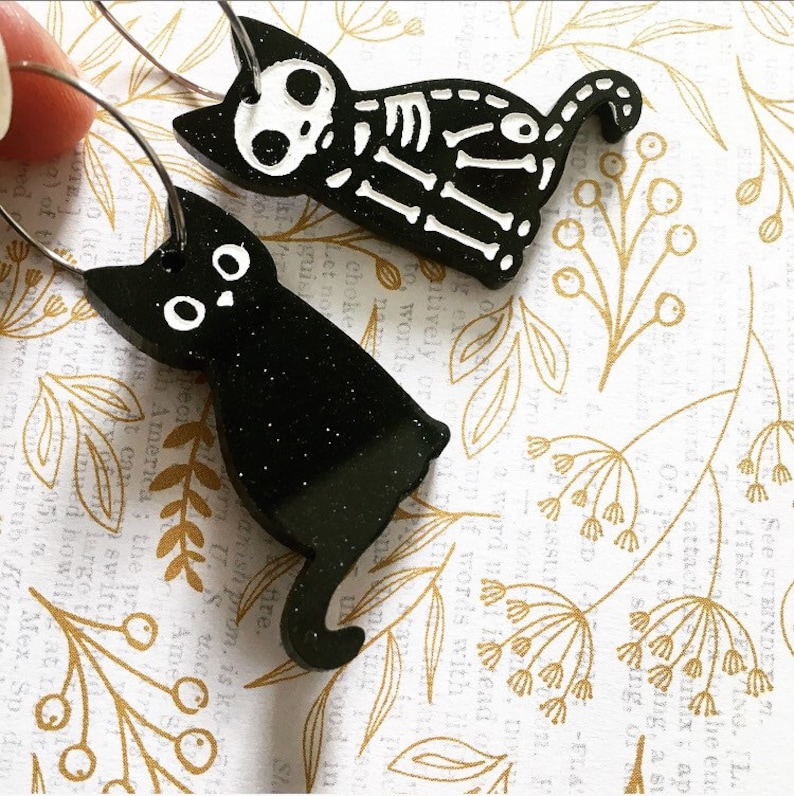 以下が含まれることがあります： キラキラ光る仕上げの黒い猫のイヤリングが2つ。左側の猫は尻尾を脚に巻き付けて座っています。右側の猫は尻尾を上げて歩いています。