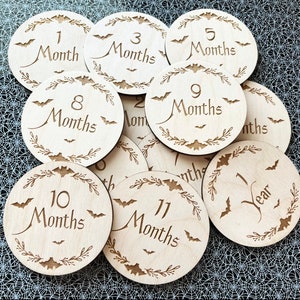 May include: A set of 11 wooden baby milestone discs with a Halloween theme. Each disc features a different number of months, from 1 to 11, with the number 12 replaced with "1 Year". The discs are decorated with bats and a leafy wreath.
