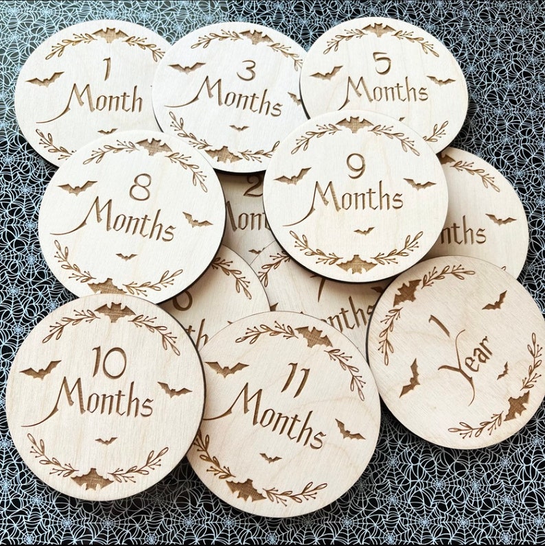 May include: A set of 11 wooden baby milestone discs with a Halloween theme. Each disc features a different number of months, from 1 to 11, with the number 12 replaced with "1 Year". The discs are decorated with bats and a leafy wreath.
