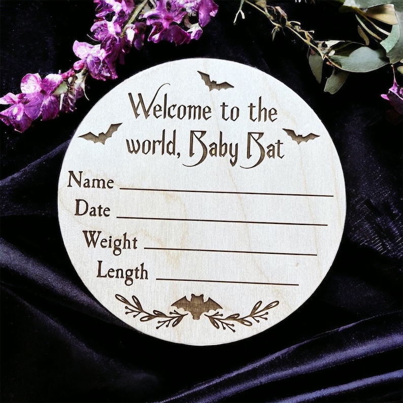May include: A round wooden sign with the text "Welcome to the world, Baby Bat" and spaces for the baby's name, date, weight, and length. The sign is decorated with bats and floral accents.