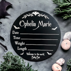 May include: Black circular wooden sign with white text and bat silhouettes. The sign reads "Ophelia Marie" and includes spaces to fill in the date, time, weight, and length. The bottom of the sign reads "Welcome to the world, Baby Bat."