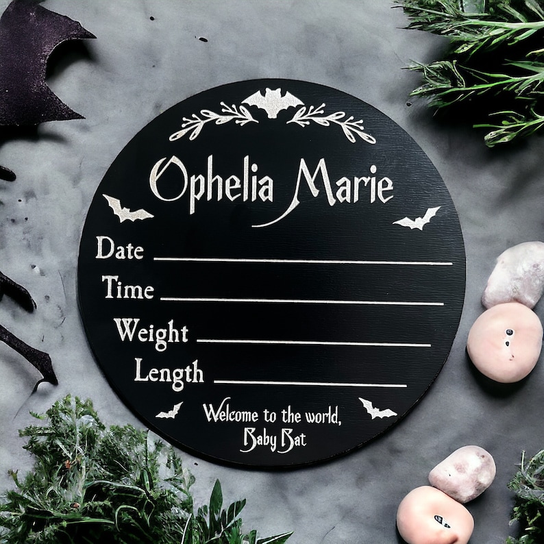 May include: Black circular wooden sign with white text and bat silhouettes. The sign reads "Ophelia Marie" and includes spaces to fill in the date, time, weight, and length. The bottom of the sign reads "Welcome to the world, Baby Bat."