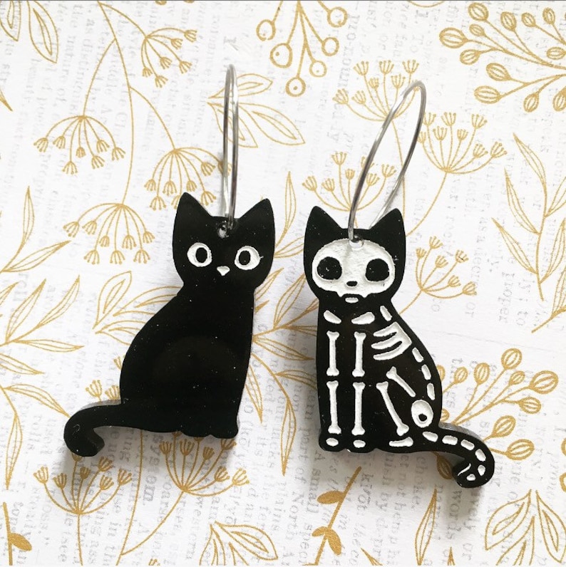 以下が含まれることがあります： 黒い猫のイヤリングのペア。1匹の猫は真っ黒で、もう1匹は白い骨のスケルトン猫です。