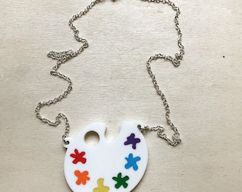 Può includere: Una collana con una tavolozza di pittura bianca e una catena argentata. La tavolozza ha un foro al centro ed è decorata con macchie di vernice colorate in tonalità di rosso, arancione, giallo, verde, blu e viola.