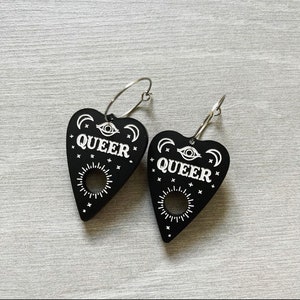 May include: A pair of black heart-shaped earrings with silver hoops. The earrings feature a white design of a crescent moon, an eye, and the word "QUEER".