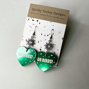 Op de afbeelding: Groene hartvormige oorbellen met de tekst "GO BIRDS!" op elke oorbel. De oorbellen hebben een zilveren stervormige hanger.