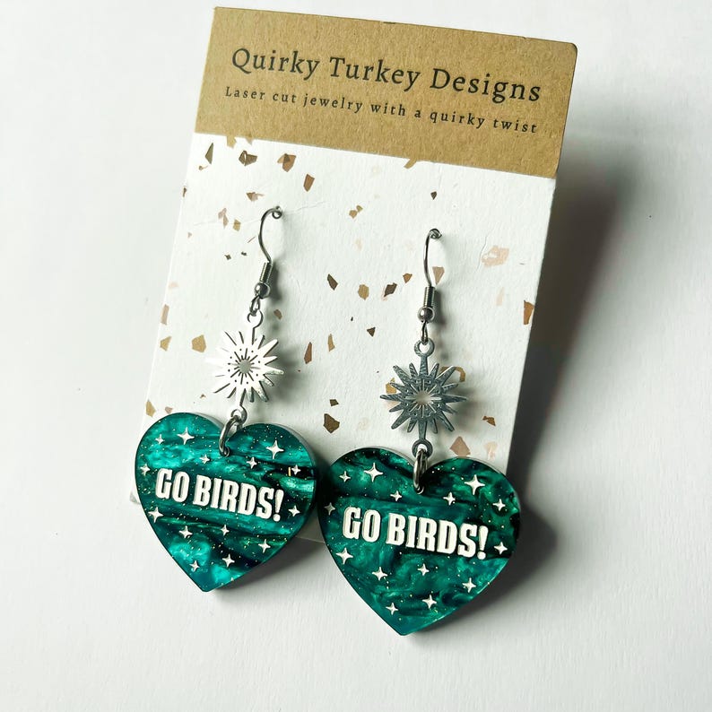 May include: A pair of green heart-shaped earrings with the text "GO BIRDS!" on each earring. The earrings are attached to silver hooks with a silver star charm.