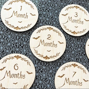 May include: Set of six wooden baby milestone discs with a Halloween theme. Each disc features a bat design and the number of months, from 1 to 7. The discs are made of natural wood and have a rustic finish.