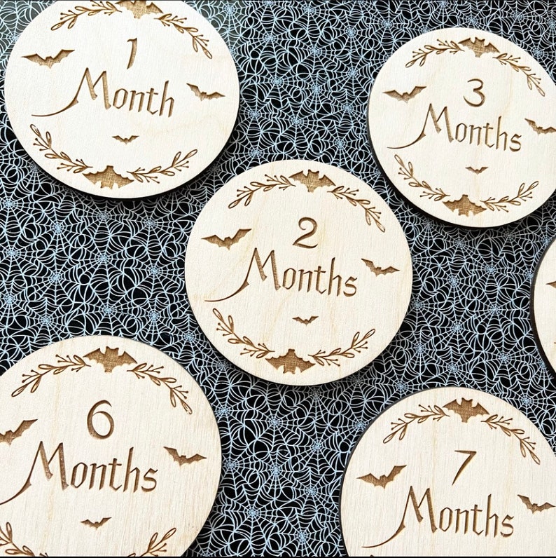 May include: Set of six wooden baby milestone discs with a Halloween theme. Each disc features a bat design and the number of months, from 1 to 7. The discs are made of natural wood and have a rustic finish.