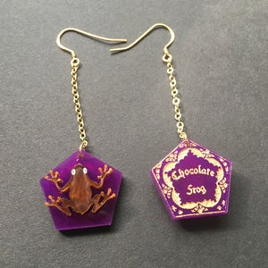 Puede incluir: Pendientes morados y dorados con un amuleto de rana de chocolate en un lado y un amuleto de caja de rana de chocolate en el otro.
