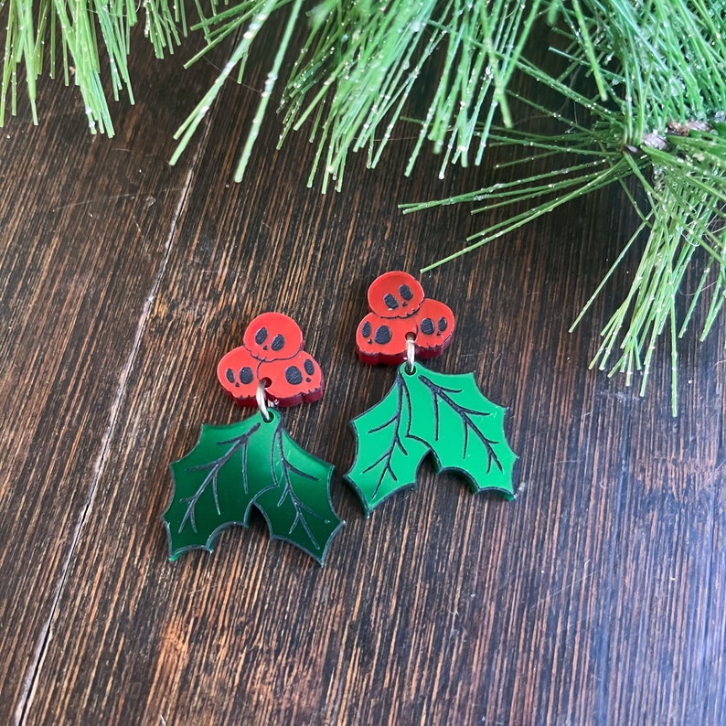 Pu&ograve; includere: Un paio di orecchini a forma di foglia di agrifoglio verde con bacche rosse a forma di teschio.