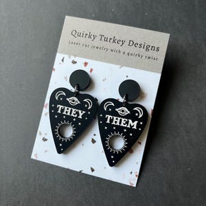 Puede incluir: Pendientes de acrílico negro en forma de tablero de Ouija de bruja con las palabras "They" y "Them" en letras blancas.