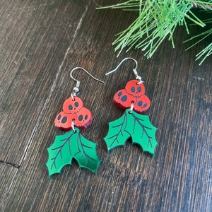 Pu&ograve; includere: Un paio di orecchini natalizi rossi e verdi con un gruppo di tre teschi rossi con occhi neri e un design a foglia di agrifoglio verde.