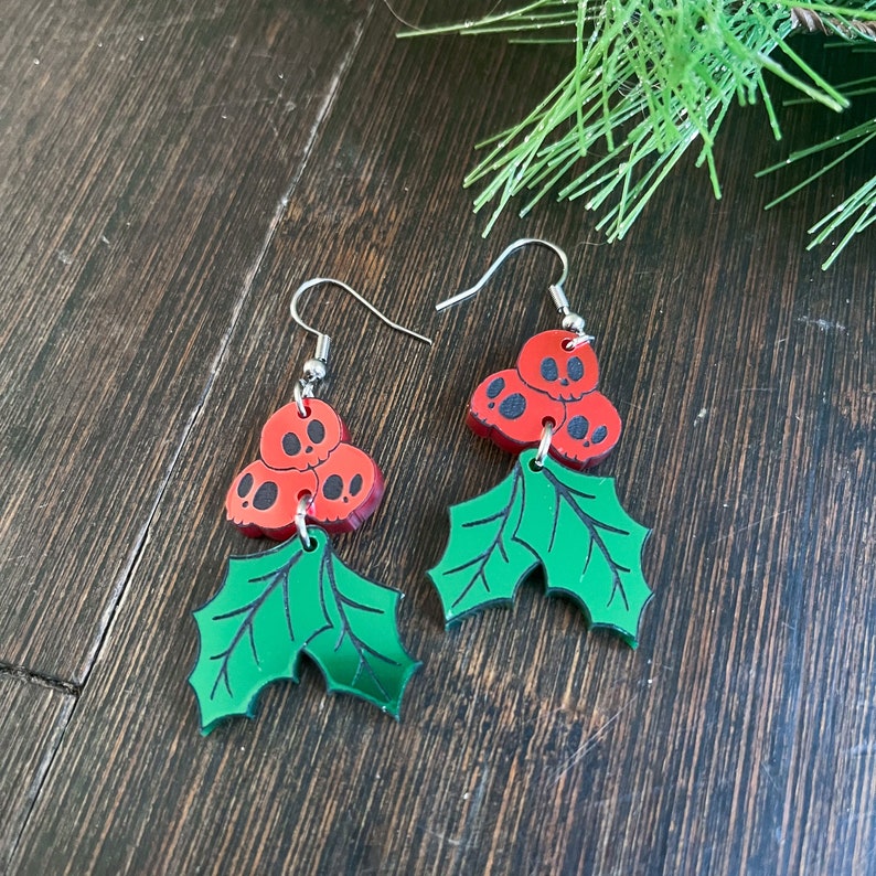 Pu&ograve; includere: Un paio di orecchini natalizi rossi e verdi con un gruppo di tre teschi rossi con occhi neri e un design a foglia di agrifoglio verde.
