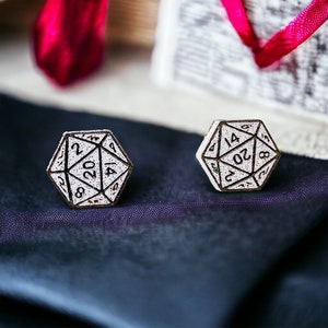 以下が含まれることがあります： 白と金色のD20サイコロ型のイヤリング2つ。各イヤリングは、白い背景に黒い数字を表示しています。イヤリングは濃い青色の布の上に置かれ、背景にはギフトボックスとリボンがぼやけています。