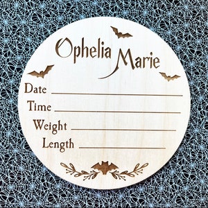 May include: A wooden round sign with a Halloween theme. It says "Ophelia Marie" and has bats and leaves carved into it. There are spaces to write the date, time, weight, and length.