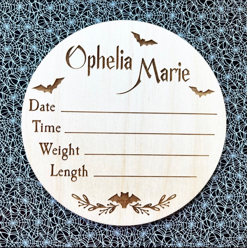 May include: A wooden round sign with a Halloween theme. It says "Ophelia Marie" and has bats and leaves carved into it. There are spaces to write the date, time, weight, and length.