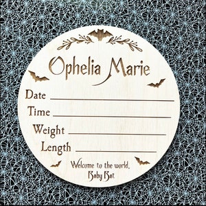 May include: A wooden baby announcement plaque with a bat and floral design. The plaque is decorated with the name "Ophelia Marie" and includes spaces to record the baby's date, time, weight, and length. The plaque also includes the text "Welcome to the world, Baby Bat."