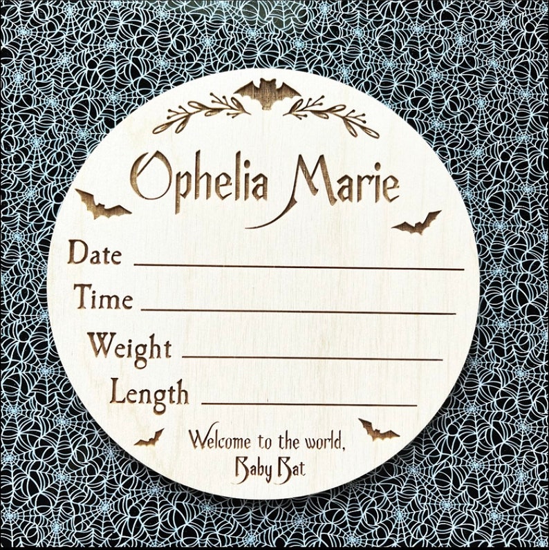 May include: A wooden baby announcement plaque with a bat and floral design. The plaque is decorated with the name "Ophelia Marie" and includes spaces to record the baby's date, time, weight, and length. The plaque also includes the text "Welcome to the world, Baby Bat."