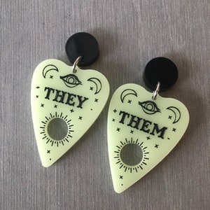 May include: A pair of glow-in-the-dark earrings shaped like a witch's board or planchette. The earrings are green and feature black text that reads "THEY" and "THEM".