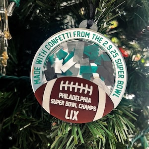 May include: A circular Christmas bauble with the text "MADE WITH CONFETTI FROM THE 2.9.25 SUPER BOWL". It features a football shape with the text "PHILADELPHIA SUPER BOWL CHAMPS LIX". The bauble is filled with confetti.