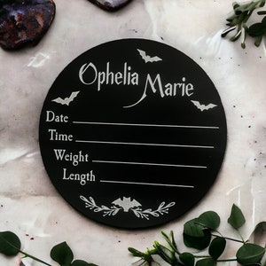 May include: Black circular wooden sign with white bats and floral accents. The sign says "Ophelia Marie" and has spaces to fill in the date, time, weight, and length.