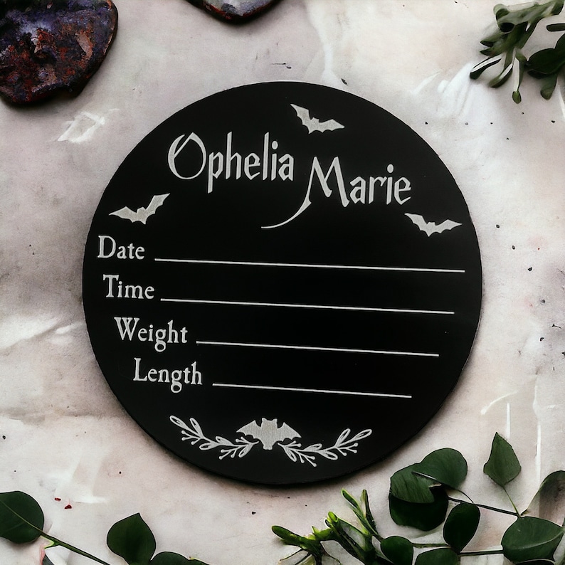 May include: Black circular wooden sign with white bats and floral accents. The sign says "Ophelia Marie" and has spaces to fill in the date, time, weight, and length.