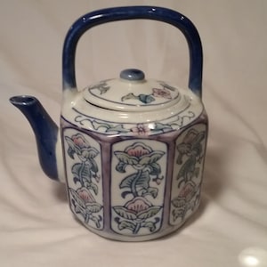 May include: A white porcelain teapot with a blue handle and lid. The teapot is decorated with floral patterns in blue, pink, and green.