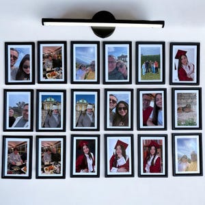 May include: A display of framed photographs on a white wall, with black frames. The images depict people and events, including graduation ceremonies and group outings. A black light fitting is positioned above the frames, illuminating the display.