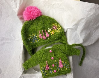 Op de afbeelding: Een groene gebreide muts en vest set met roze pompon en geborduurde bloemen.