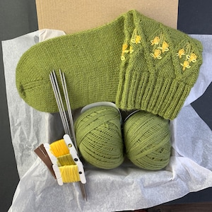 Könnte beinhalten: Eine grüne Stricksocke mit gelben gestickten Blumen, zwei Knäuel grünes Garn und Stricknadeln.