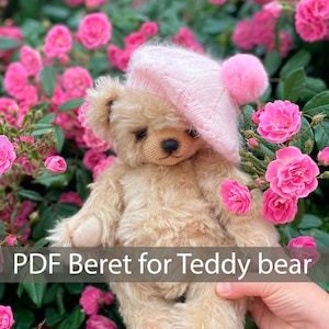 May include: A beige teddy bear wearing a pink knitted beret with a pom-pom. The bear is sitting in a hand against a background of pink roses.