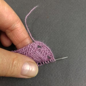 May include: A close-up of a purple knitted fabric with a needle threaded through it. The fabric has a lacy pattern and is being held by a hand.