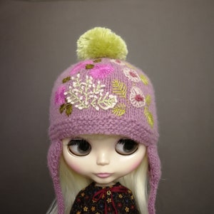 May include: A purple knitted hat with a green pom pom and embroidered flowers. The hat has ear flaps and is designed for a doll.