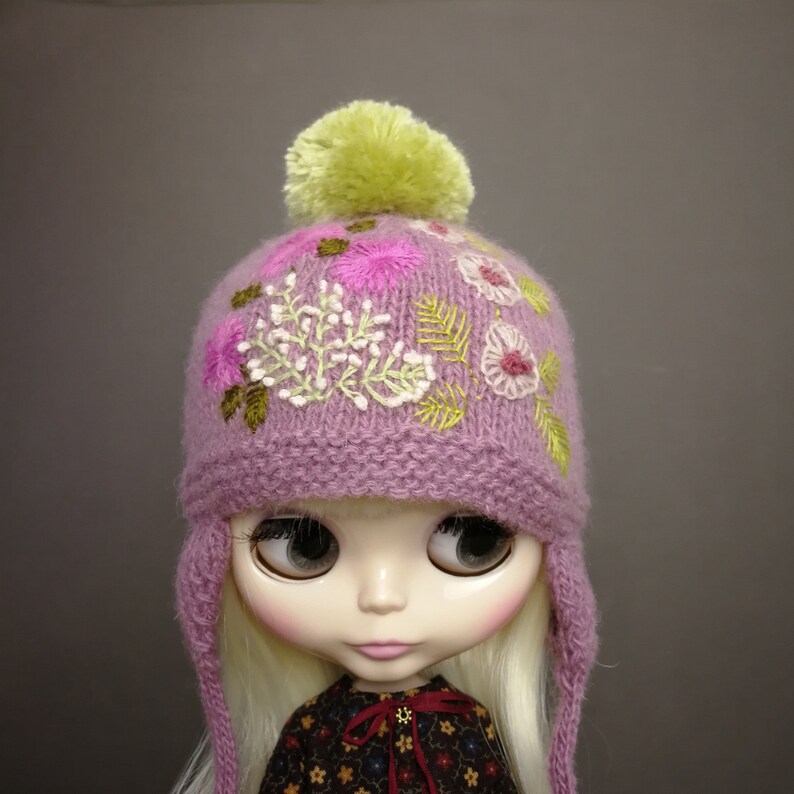 May include: A purple knitted hat with a green pom pom and embroidered flowers. The hat has ear flaps and is designed for a doll.