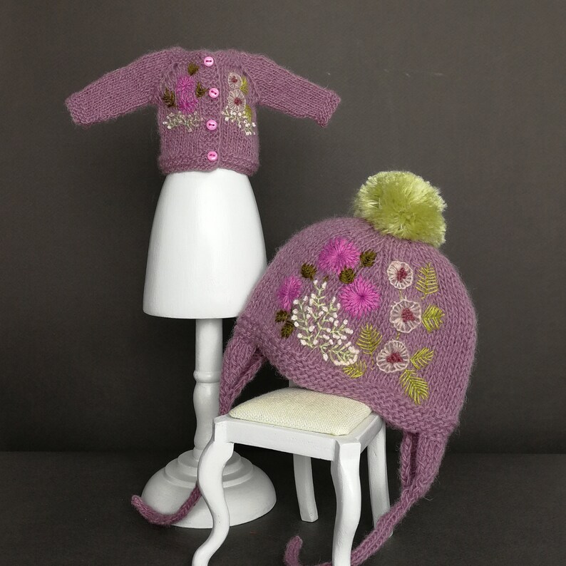 May include: A set of knitted baby clothes in a mauve color with floral embroidery. The set includes a cardigan and a hat with a pom-pom.
