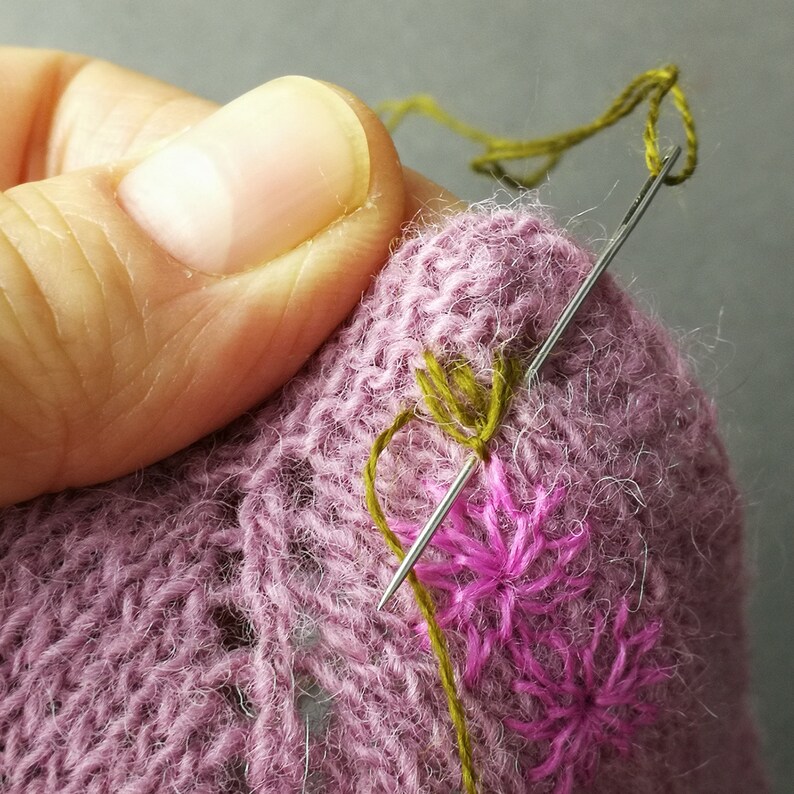 May include: Close-up of a hand embroidering pink flowers on a purple knitted fabric with green thread. The needle and thread are visible.
