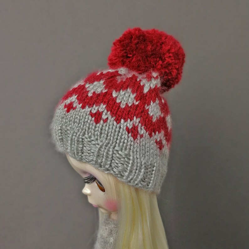 May include: A gray and red knitted hat with a pom-pom on top. The hat has a fair isle pattern with red and white stars.