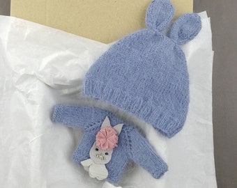 Op de afbeelding: Een blauwe gebreide muts met konijnenoren en een bijpassende blauwe gebreide trui met een roze bloem en een grijze "love" applicatie. De items zijn verpakt in wit tissuepapier.
