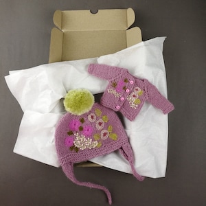 May include: A pink knitted hat and cardigan set with floral embroidery. The hat has a pom-pom on top and the cardigan has button closures.