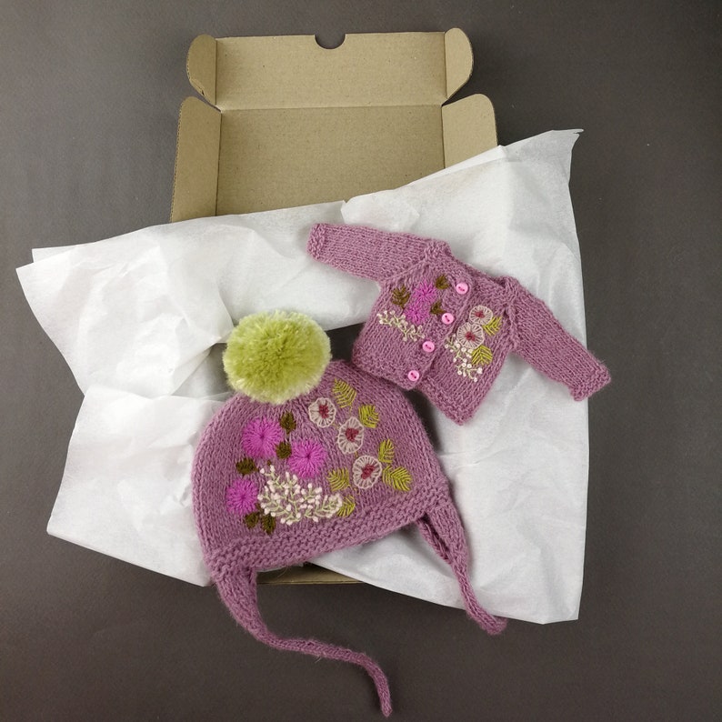 May include: A pink knitted hat and cardigan set with floral embroidery. The hat has a pom-pom on top and the cardigan has button closures.
