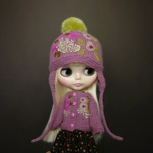 May include: A doll wearing a pink knitted hat with embroidered flowers and a matching pink cardigan. The hat has a green pom-pom on top.