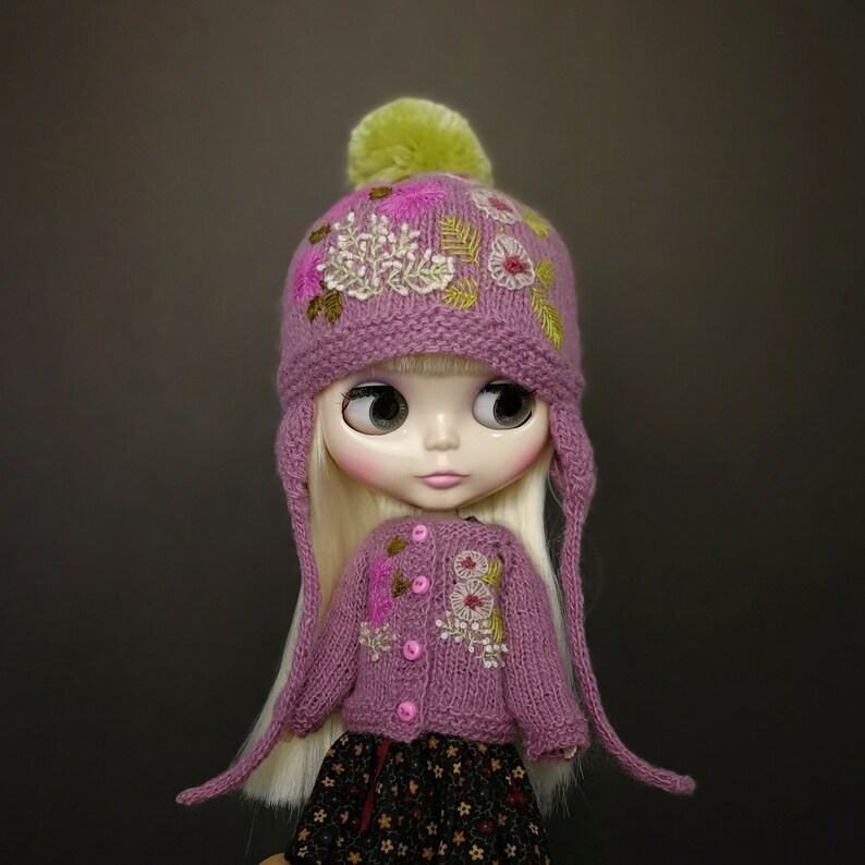 May include: A doll wearing a pink knitted hat with embroidered flowers and a matching pink cardigan. The hat has a green pom-pom on top.
