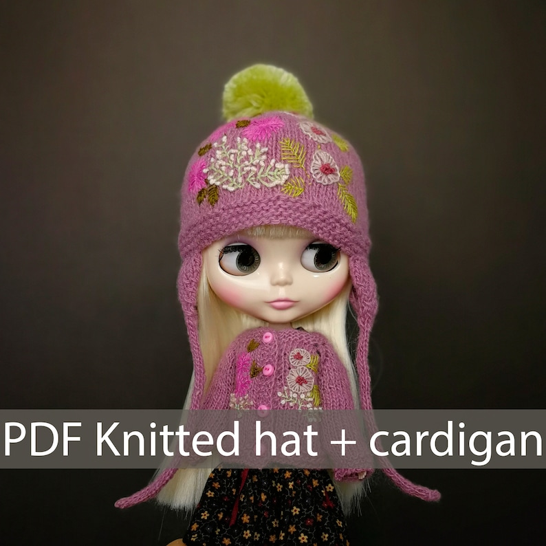 May include: A pink knitted hat with a green pom pom and floral embroidery, paired with a matching pink cardigan with floral embroidery. The hat and cardigan are designed for a doll.